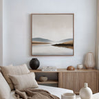 A minimalistic, cozy living room features a beige sofa, neutral pillows, a wooden sideboard, decorative vases, stacked books, and is completed by the Tranquil Waters Canvas Art - Square for a serene lakeside-inspired decor.