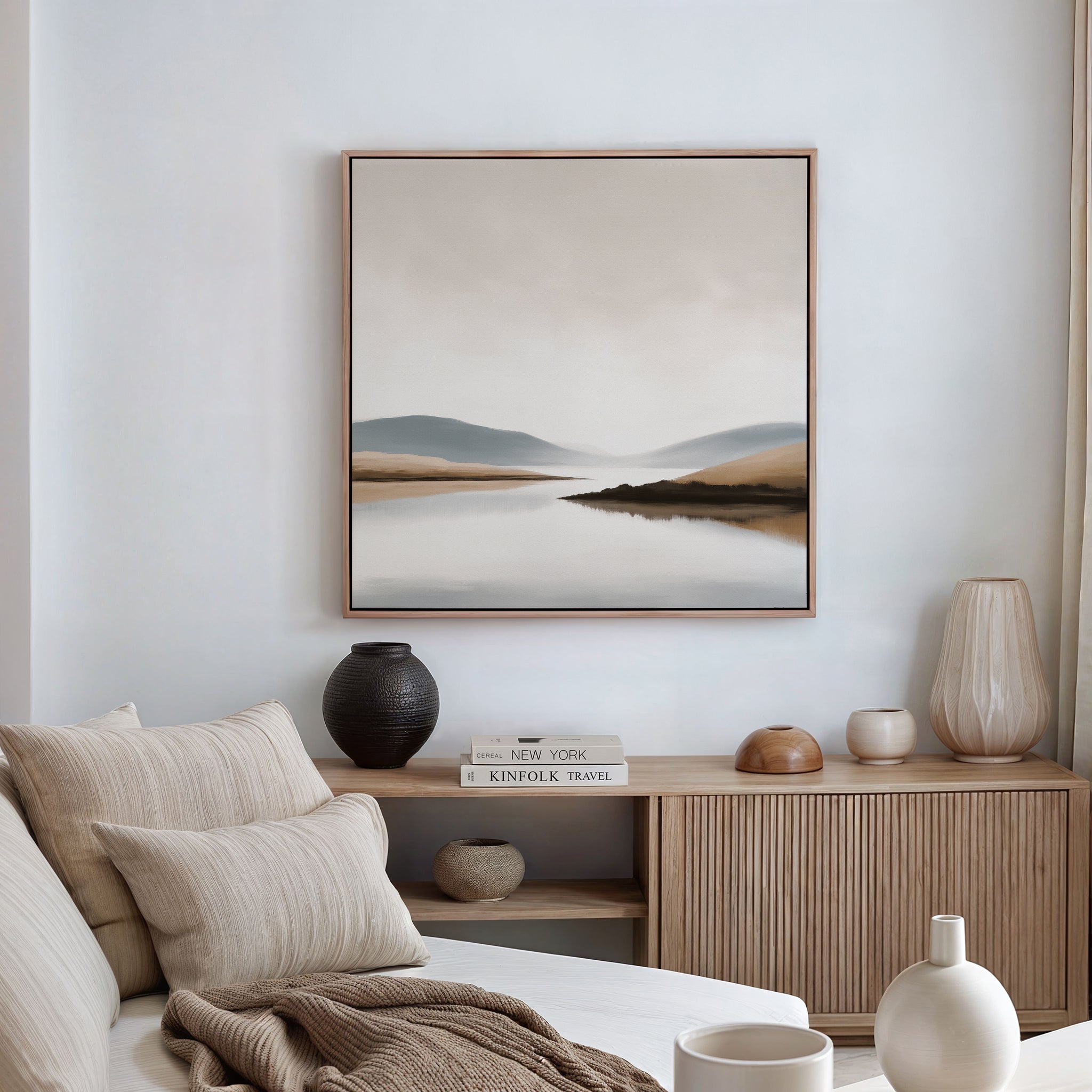 A minimalistic, cozy living room features a beige sofa, neutral pillows, a wooden sideboard, decorative vases, stacked books, and is completed by the Tranquil Waters Canvas Art - Square for a serene lakeside-inspired decor.