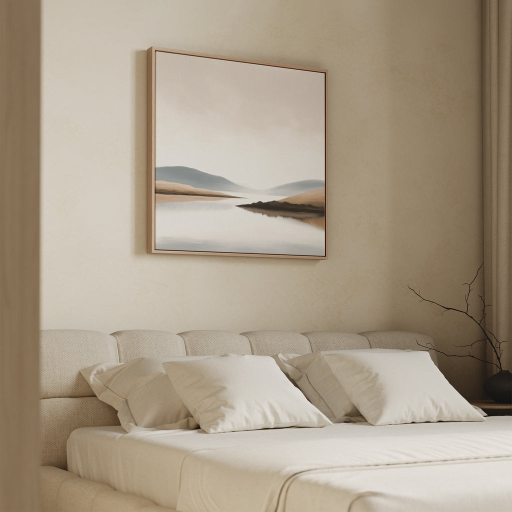 A modern bedroom features a beige upholstered bed with white bedding, and the Tranquil Waters Canvas Art - Square above the headboard. A dry branch in a vase adds a natural touch to the serene lakeside setting.
