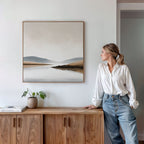A woman in a white blouse and jeans leans on a wooden cabinet, admiring the Tranquil Waters Canvas Art - Square—ideal for serene living room decor. A plant and book add simple touches beside her.