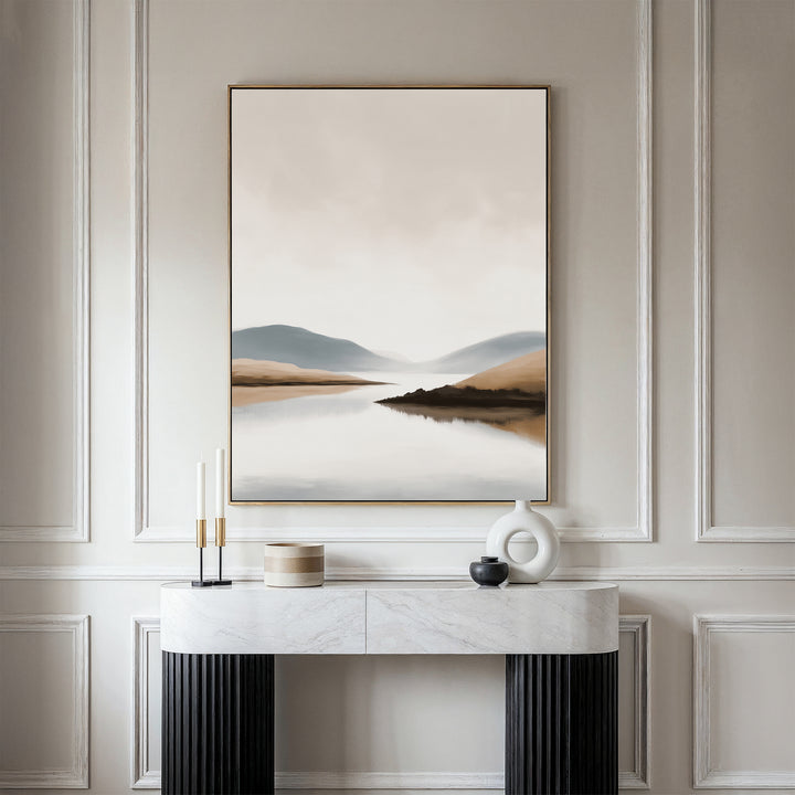 A minimal console table with decor sits against a white paneled wall, topped by the Tranquil Waters Canvas Art - Vertical—a serene landscape featuring misty mountains and calm lakeside views for a soothing touch.