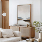A minimalist living room with a light wooden cabinet, neutral-toned decor, the Tranquil Waters Canvas Art - Vertical above a white sofa with a cream pillow, a round lamp, and a small leafy plant on a white table.