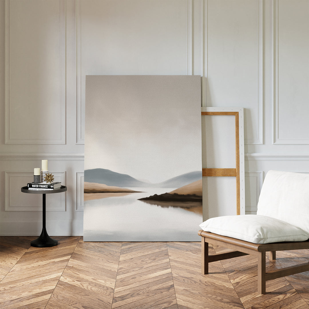 A minimalist living room showcases the Tranquil Waters Canvas Art - Vertical, featuring a serene mountain and lake scene, propped against a white paneled wall beside a white cushioned chair and black side table on herringbone wood flooring.