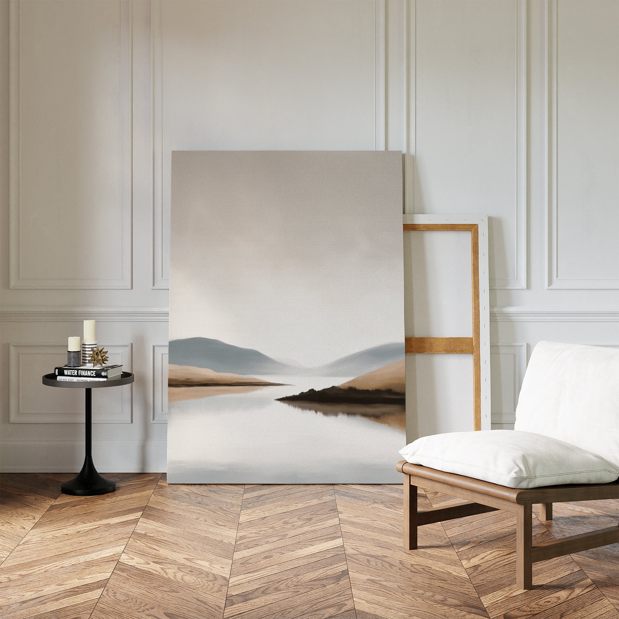 A minimalist living room showcases the Tranquil Waters Canvas Art - Vertical, featuring a serene mountain and lake scene, propped against a white paneled wall beside a white cushioned chair and black side table on herringbone wood flooring.