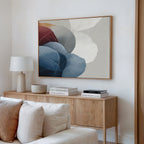 A modern living room corner with a wooden sideboard, white lamp, stacked books, a ceramic bowl, and Translucent Petals Canvas Art - Horizontal—an abstract painting featuring blue, maroon, and beige shapes—displayed above.