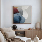 A modern living room with a beige sofa, neutral cushions, a wooden sideboard, and ceramics. The Translucent Petals Canvas Art - Square, featuring blue, red, and white shapes, hangs above the sideboard.