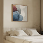A minimalistic bedroom with a beige upholstered bed, white bedding, and neutral pillows. Above the bed hangs the Translucent Petals Canvas Art - Square in blue, red, and light tones. A bare branch decorates a vase on a side table.