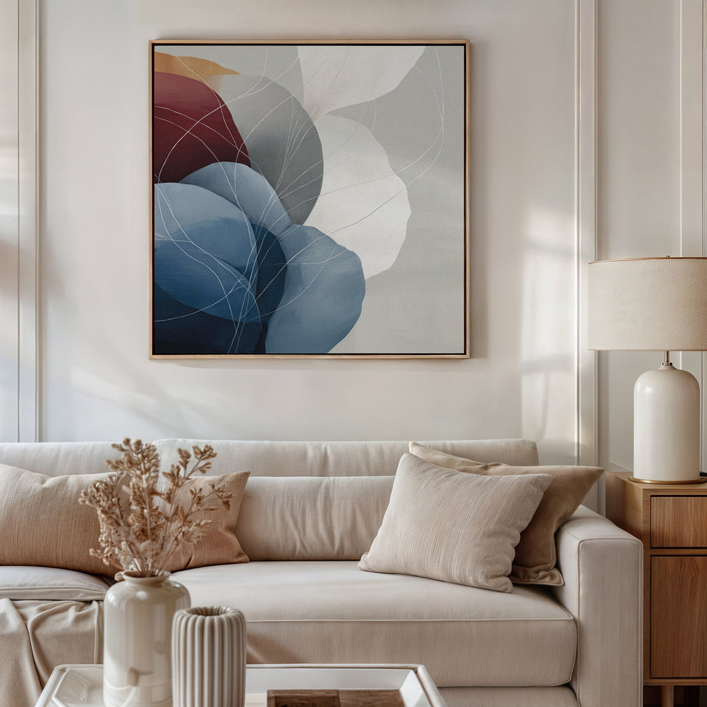 A modern living room with a beige sofa, neutral cushions, dried flowers in a vase, a table lamp on the coffee table, and the Translucent Petals Canvas Art - Square featuring blue, burgundy, and white shapes hanging on the wall.