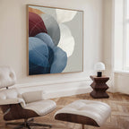 A modern living room with a white leather lounge chair and ottoman, a geometric wood side table with a small white lamp, and the Translucent Petals Canvas Art - Square featuring soft blue, burgundy, and white shapes on the wall.