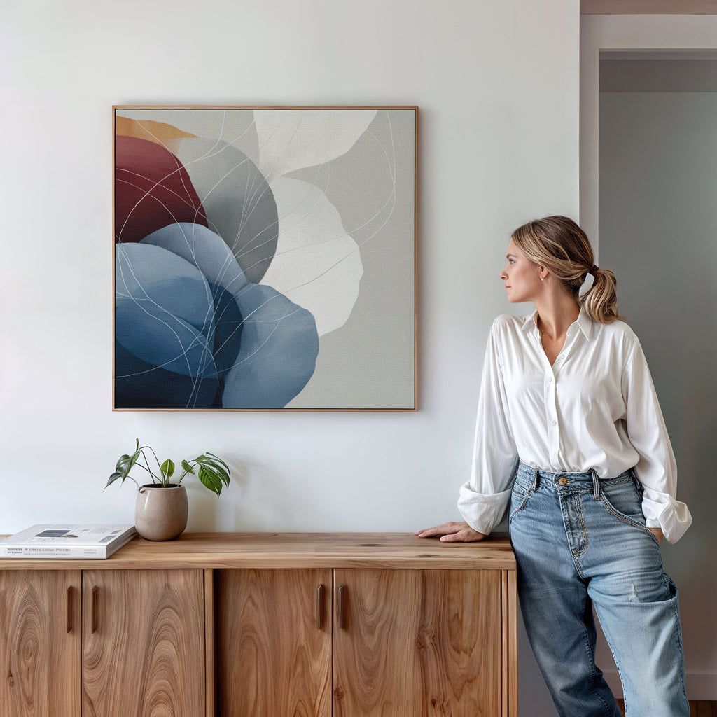 A woman in a white blouse and jeans leans against a wooden sideboard, admiring the Translucent Petals Canvas Art - Square, which displays blue, burgundy, and neutral organic shapes on a white wall.
