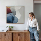 A woman in a white blouse and jeans leans against a wooden sideboard, admiring the Translucent Petals Canvas Art - Square, which displays blue, burgundy, and neutral organic shapes on a white wall.