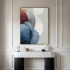 A modern console table with decorative items sits below the Translucent Petals Canvas Art - Vertical, featuring overlapping blue, red, and white shapes, all set against an elegant cream-paneled wall.