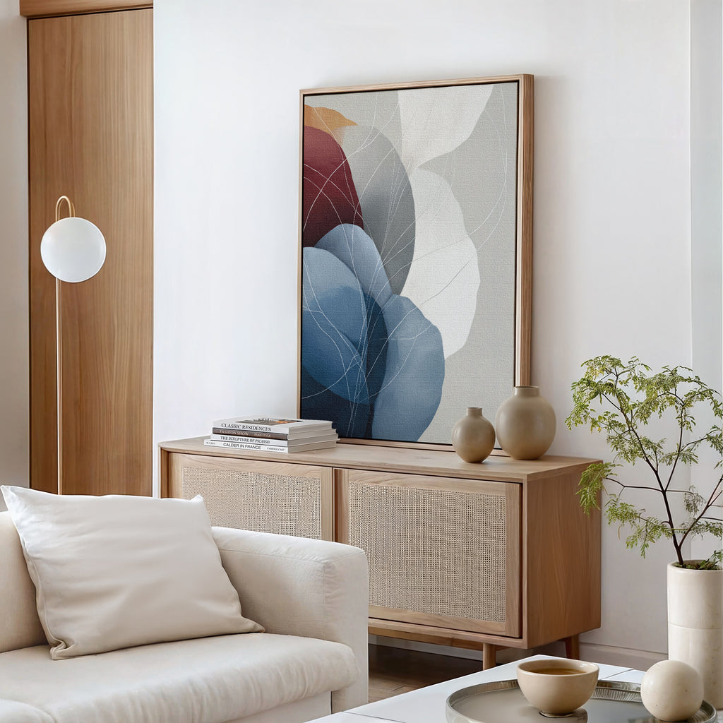 A modern, minimalist living room with a light wood sideboard, Translucent Petals Canvas Art - Vertical in blue, beige, and red tones, ceramic vases, a small green plant, and a cream sofa with cushions. The decor feels neutral and calming.