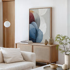 A modern, minimalist living room with a light wood sideboard, Translucent Petals Canvas Art - Vertical in blue, beige, and red tones, ceramic vases, a small green plant, and a cream sofa with cushions. The decor feels neutral and calming.