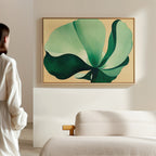 A person in a white shirt stands in a bright, minimal room, admiring the Tropical Serenity Canvas Art - Horizontal featuring green leaves on a beige wall above a soft sofa—perfect for lovers of neutral wall decor.