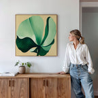 A woman in a white blouse and jeans stands by a wooden sideboard, admiring the Tropical Serenity Canvas Art - Square, featuring a large green and cream abstract leaf. A small potted plant and books complete the serene, beachy atmosphere.