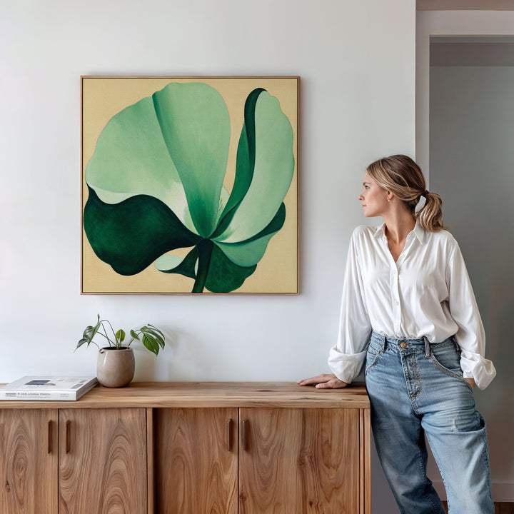A woman in a white blouse and jeans stands by a wooden sideboard, admiring the Tropical Serenity Canvas Art - Square, featuring a large green and cream abstract leaf. A small potted plant and books complete the serene, beachy atmosphere.