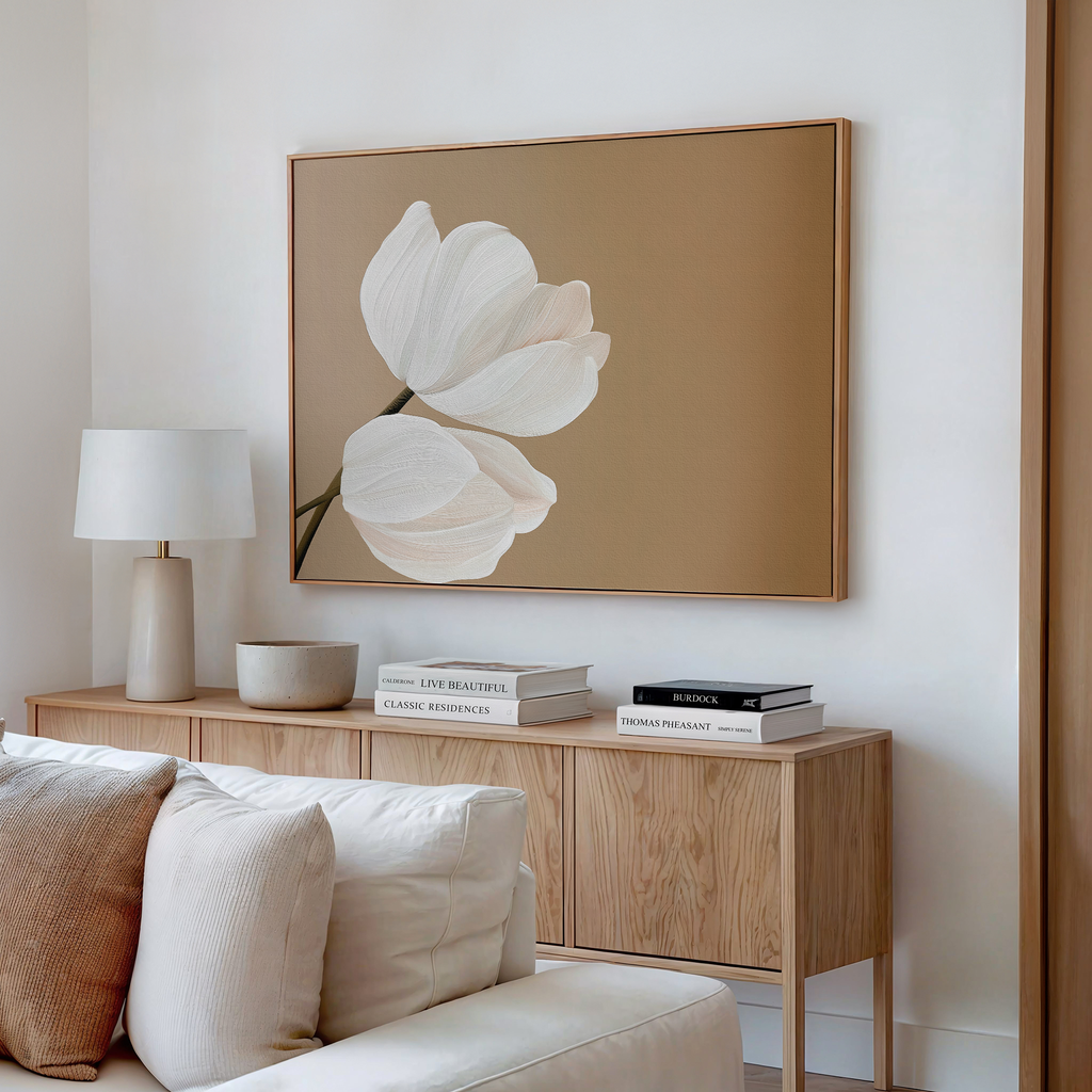 A modern living room features a light wood sideboard, white lamp, books, a bowl, beige couch with cream pillows, and Tulips Abstract Canvas Art—large minimalist wall art with two white flowers on a tan background.