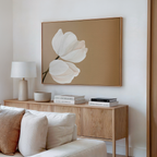 A modern living room features a light wood sideboard, white lamp, books, a bowl, beige couch with cream pillows, and Tulips Abstract Canvas Art—large minimalist wall art with two white flowers on a tan background.