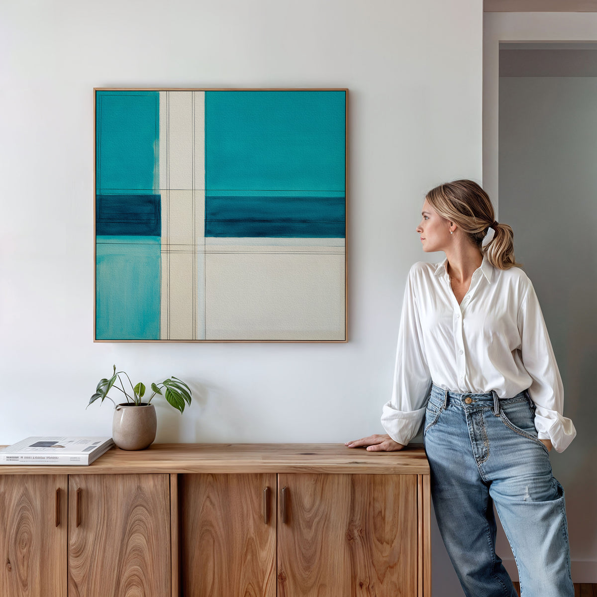 A woman in a white blouse and jeans leans against a wooden cabinet, admiring the Turquoise Grid Canvas Art - Square—a modern abstract piece with bold blue and teal blocks—hanging above, while a small plant and books sit on the cabinet.
