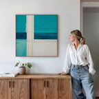 A woman in a white blouse and jeans leans against a wooden cabinet, admiring the Turquoise Grid Canvas Art - Square—a modern abstract piece with bold blue and teal blocks—hanging above, while a small plant and books sit on the cabinet.