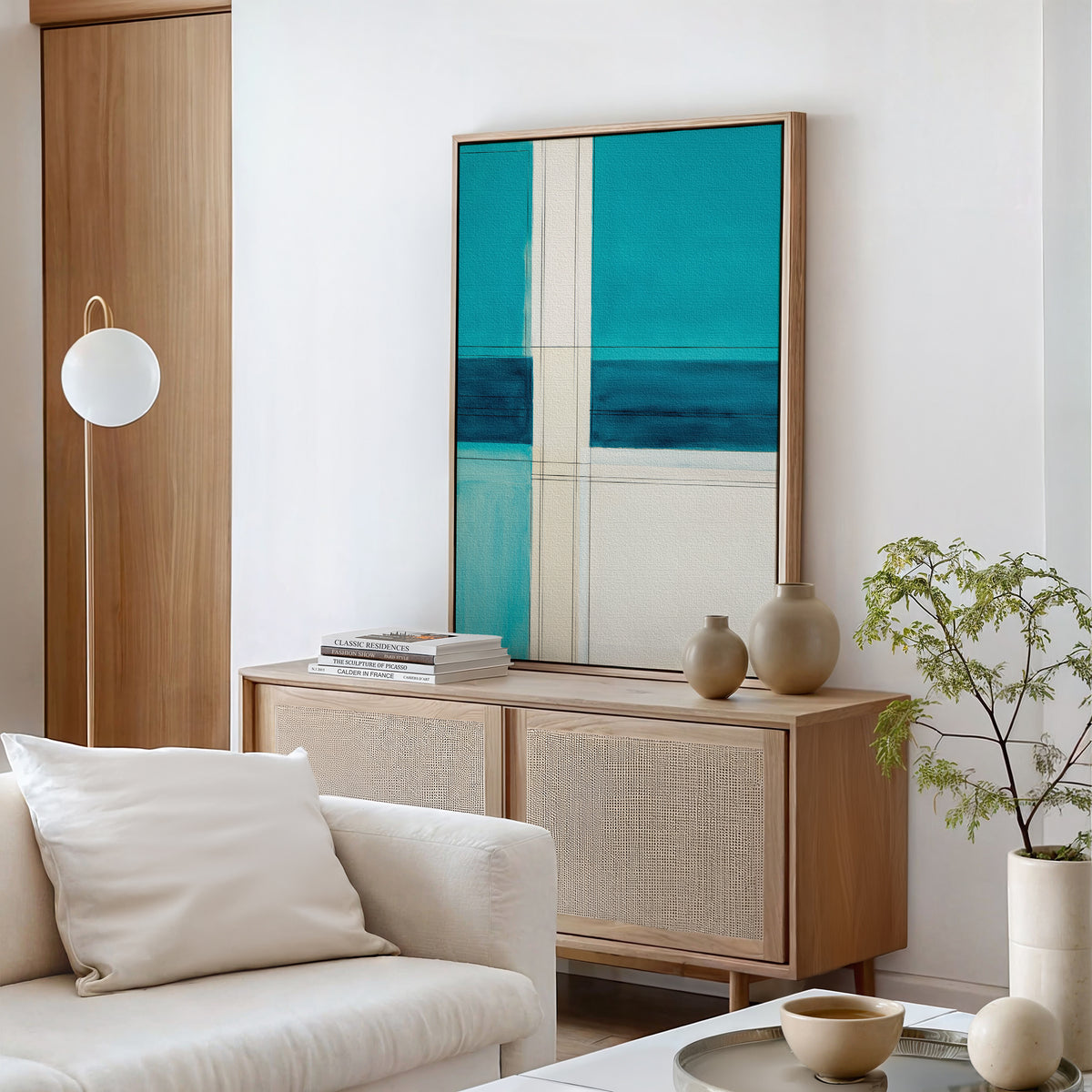 A modern living room with a light wood sideboard, Turquoise Grid Canvas Art - Vertical above it, ceramic vases, stacked books, white sofa and cushion, round lamp, and a small potted plant.