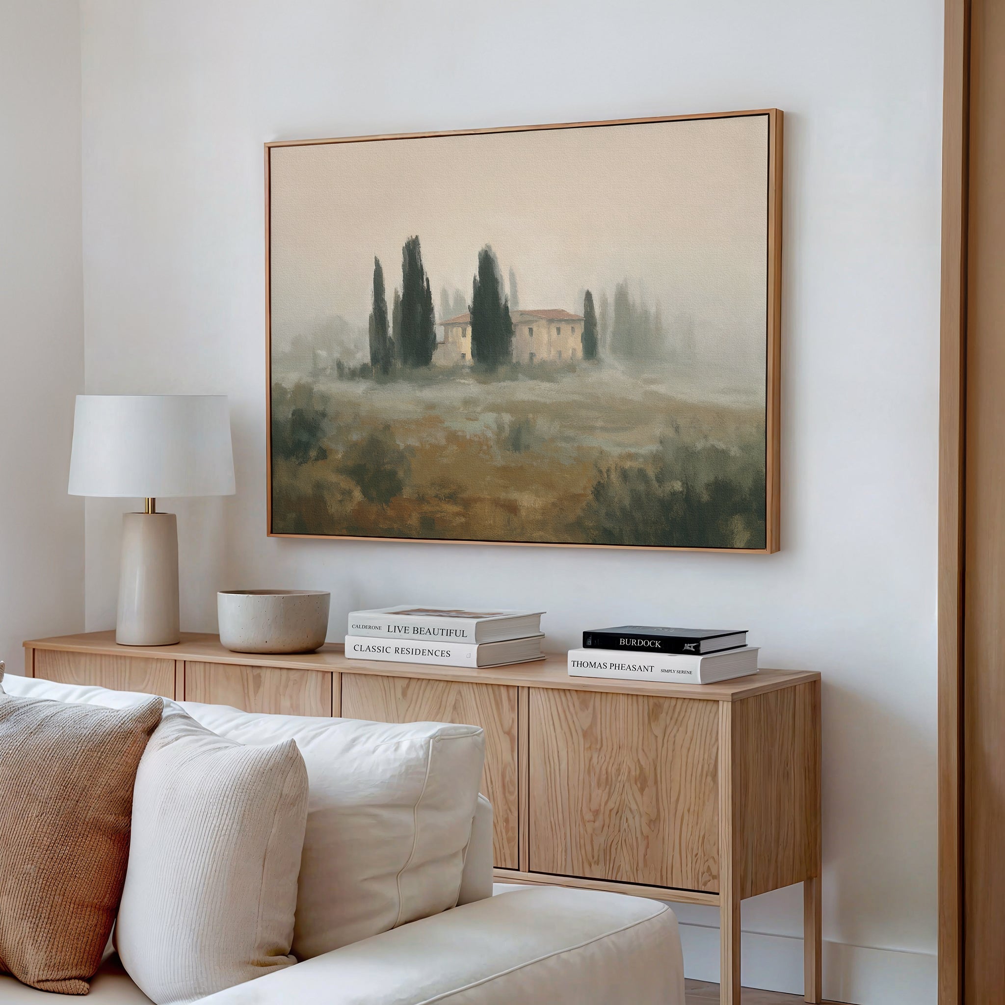 A modern living room features a light wood console, beige sofa, white lamp, stacked books, and decor. Above the console hangs the Tuscan Veil Canvas Art - Horizontal, showcasing a Mediterranean landscape with a country house and trees.