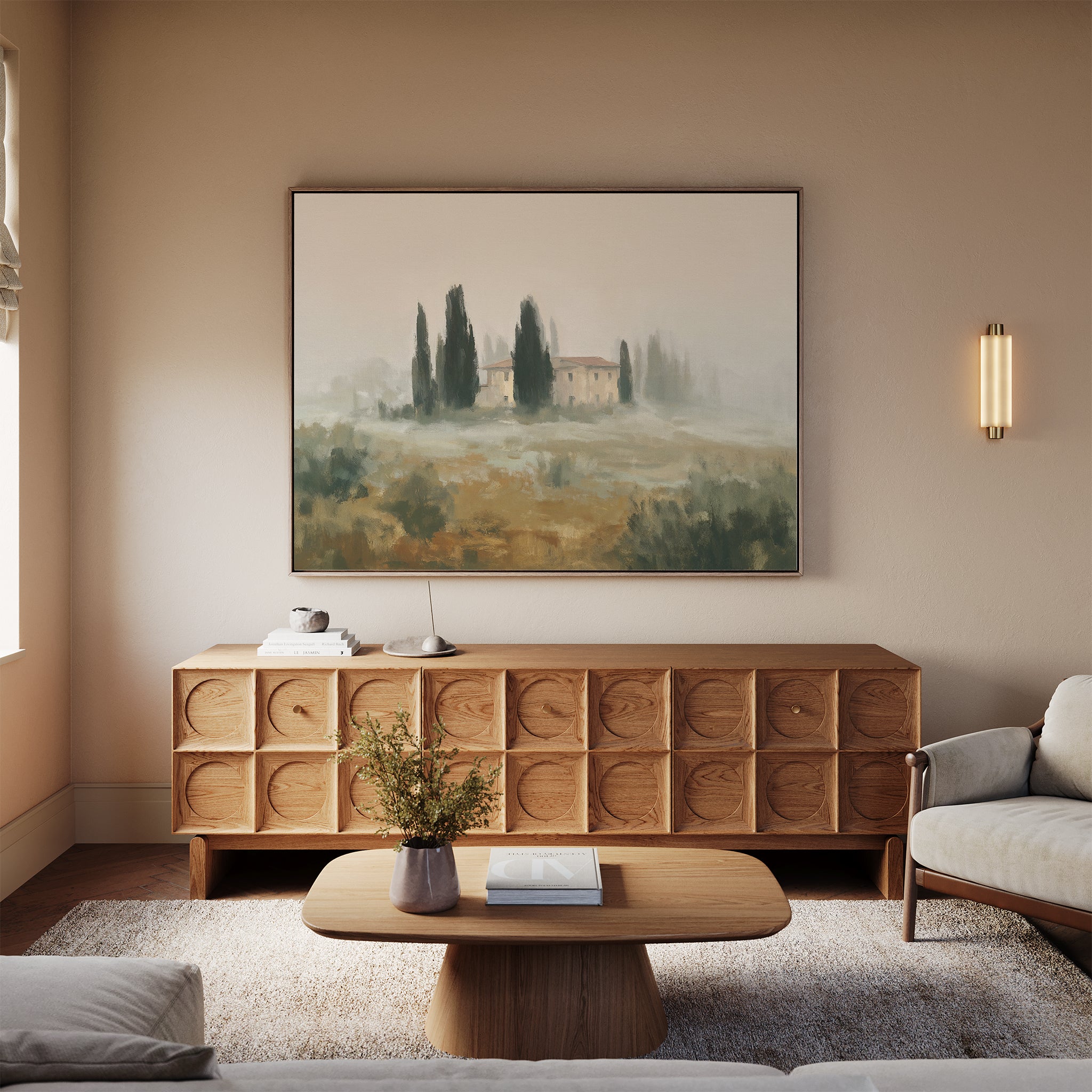 A cozy living room with a light sofa, wooden coffee table, and round sideboard features the Tuscan Veil Canvas Art - Horizontal. A landscape painting and small lamp brighten the neutral space, with a vase of flowers adding charm to the table.