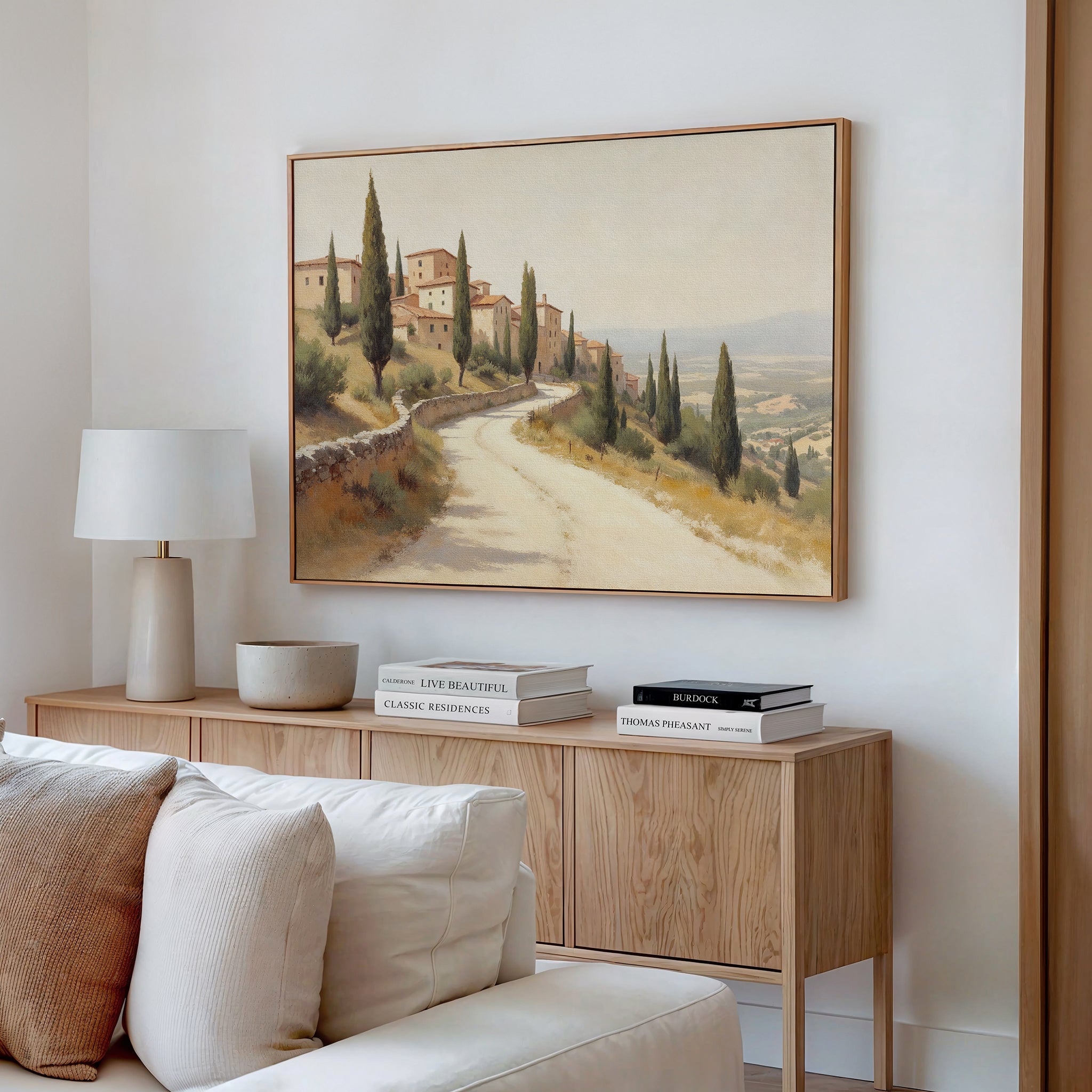 A cozy living room corner features a light wood console table with books, a lamp, and beige decor accents. Above the table, the Tuscan Village Canvas Art - Horizontal depicts a country road with cypress trees on a white wall.