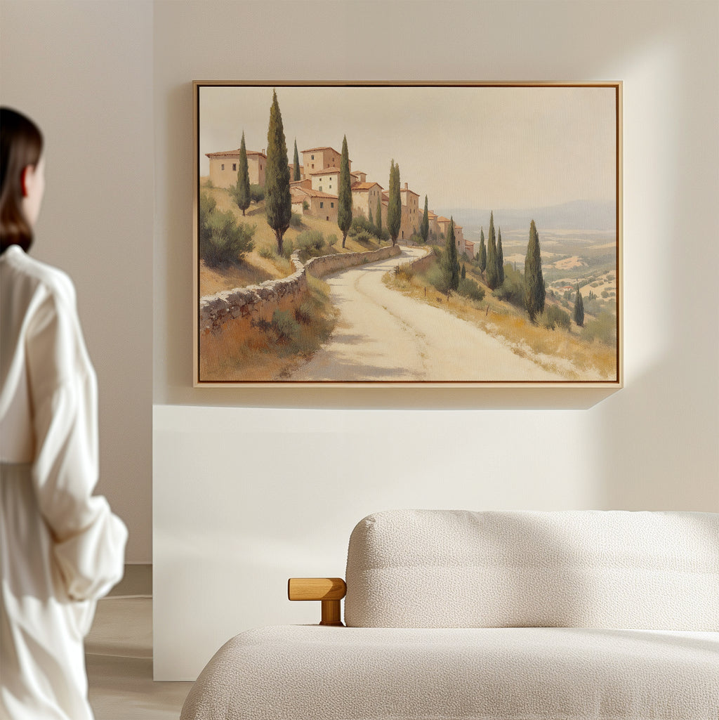 A person stands in a softly lit room, gazing at the Tuscan Village Canvas Art - Horizontal—a rustic landscape of a winding road past trees and buildings—hanging above a light upholstered sofa on a neutral wall.