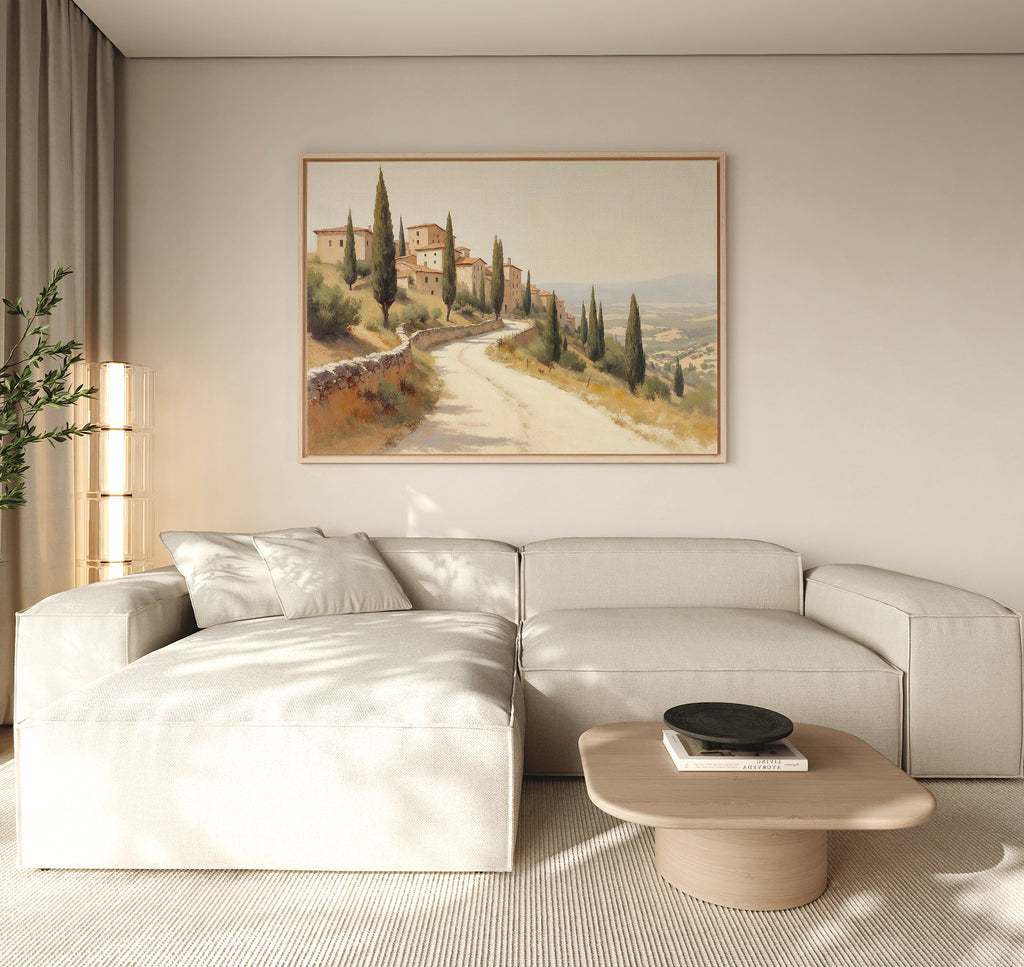 A modern living room with a light beige sectional sofa, a low wooden coffee table with a black dish, neutral walls, and rustic wall art featuring the Tuscan Village Canvas Art - Horizontal depicting a countryside road with trees and houses.