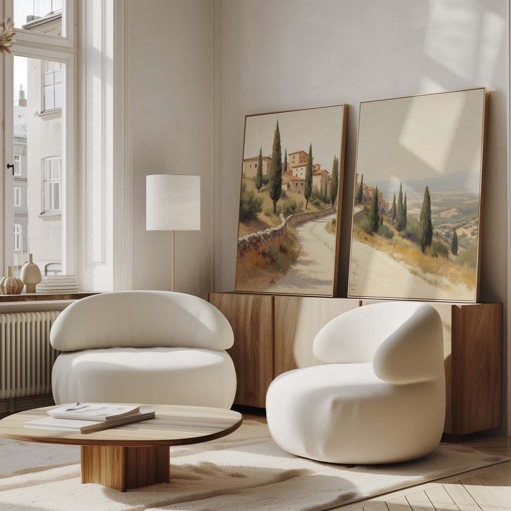 A modern living room features two curved white chairs, a round wooden coffee table, a wooden sideboard, and the Tuscan Village - Set of 2 canvas prints leaning against the wall near a tall window.