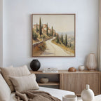 A neutral-toned living room with a beige sofa, striped pillows, and a wooden sideboard is accented by the Tuscan Village Canvas Art - Square above decorative vases and stacked books.