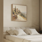 A cozy bedroom features a beige bed with white pillows, neutral walls, and the Tuscan Village Canvas Art - Square above the bed, showcasing a Mediterranean countryside road lined with cypress trees and rustic buildings.