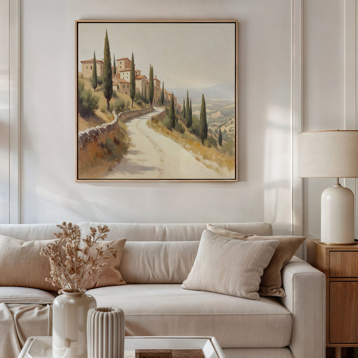A neutral-toned living room with a beige sofa, cream pillows, a side table with a lamp, and dried flowers. Above the sofa hangs the Tuscan Village Canvas Art - Square, depicting a countryside road lined with cypress trees.