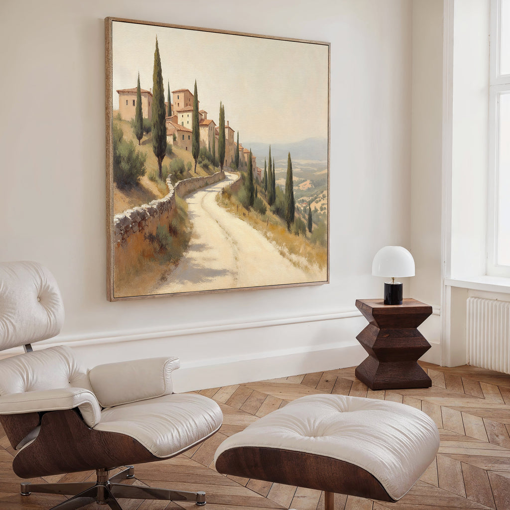 A modern living room with a white lounge chair and ottoman, dark wooden side table with a white lamp, and the Tuscan Village Canvas Art - Square featuring a sunlit country road lined with cypress trees and village buildings.