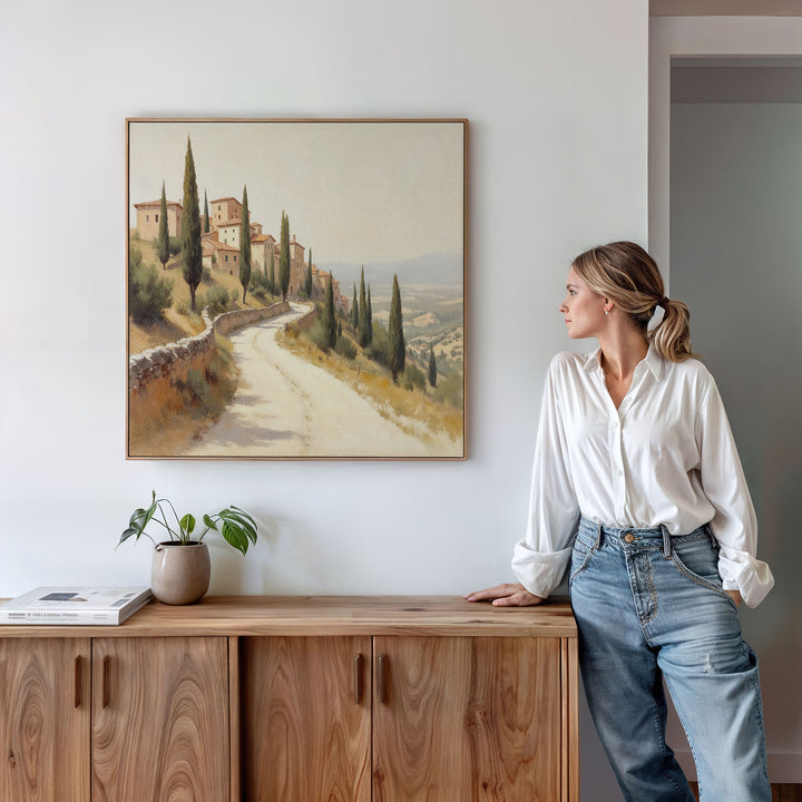 A woman in a white blouse and jeans leans on a wooden cabinet, gazing at the Tuscan Village Canvas Art - Square—a rustic landscape of countryside houses—hanging on a white wall. A small plant and book rest on the cabinet.