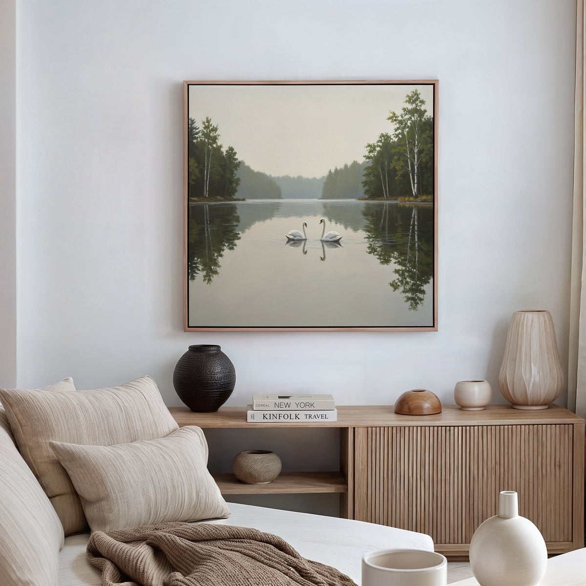 A cozy living room features a beige sofa, neutral pillows, a wooden sideboard with vases and books, and above, the Twin Swans Serenity Canvas Art—a square painting of two swans on a calm lake in Nordic Park.