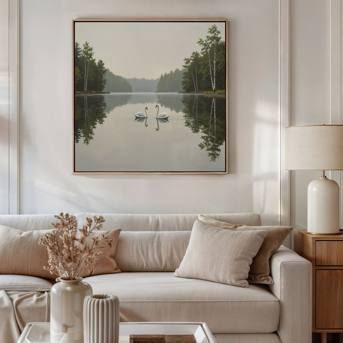 A cozy living room features a light sofa, beige pillows, and a table lamp. On the wall, Twin Swans Serenity Canvas Art - Square—a painting of two swans on a calm lake—adds a peaceful touch to the space.