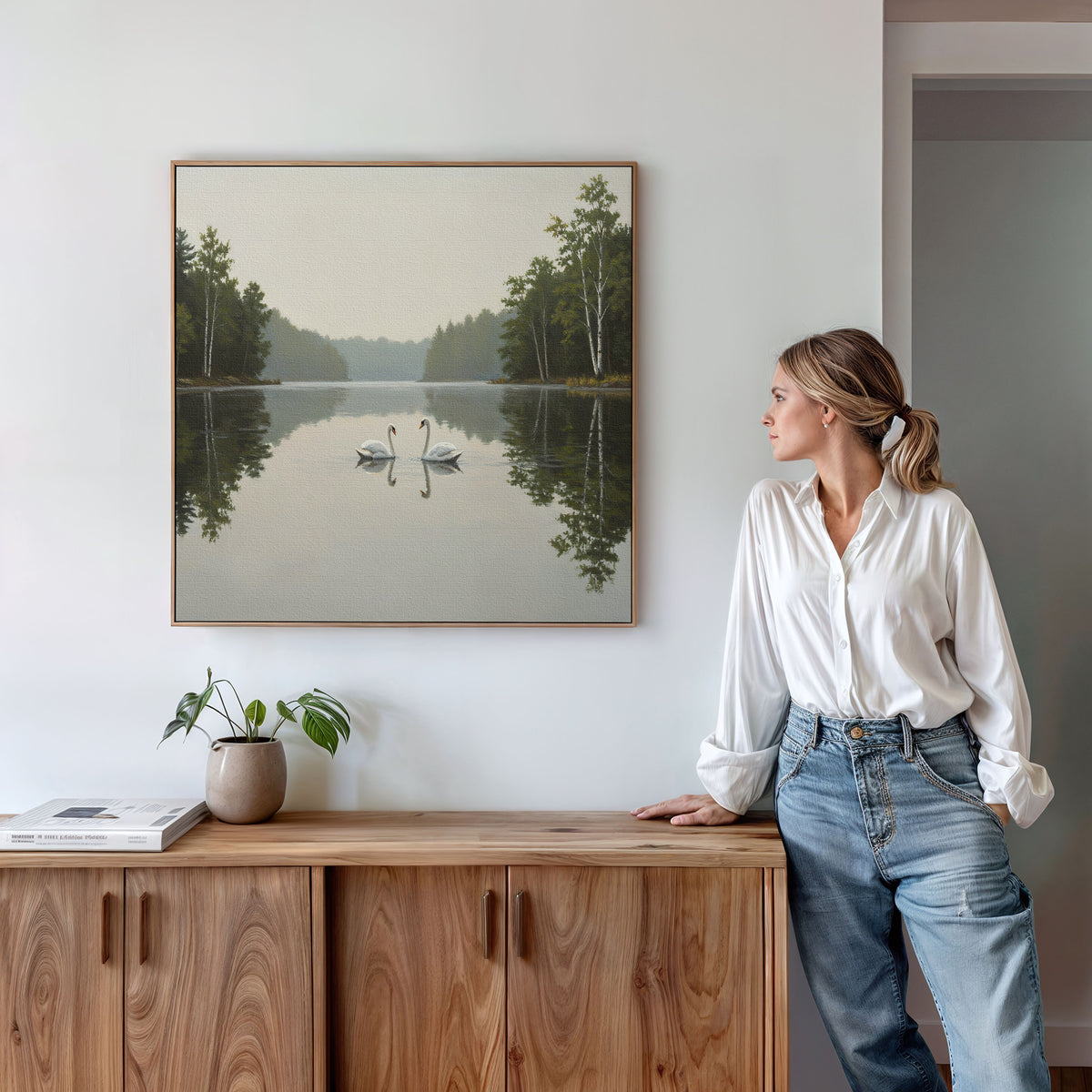 A woman in a white blouse and jeans stands by a wooden sideboard, admiring the Twin Swans Serenity Canvas Art - Square, which features serene swans with calm waters and trees, hanging on a white wall.