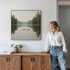 A woman in a white blouse and jeans stands by a wooden sideboard, admiring the Twin Swans Serenity Canvas Art - Square, which features serene swans with calm waters and trees, hanging on a white wall.