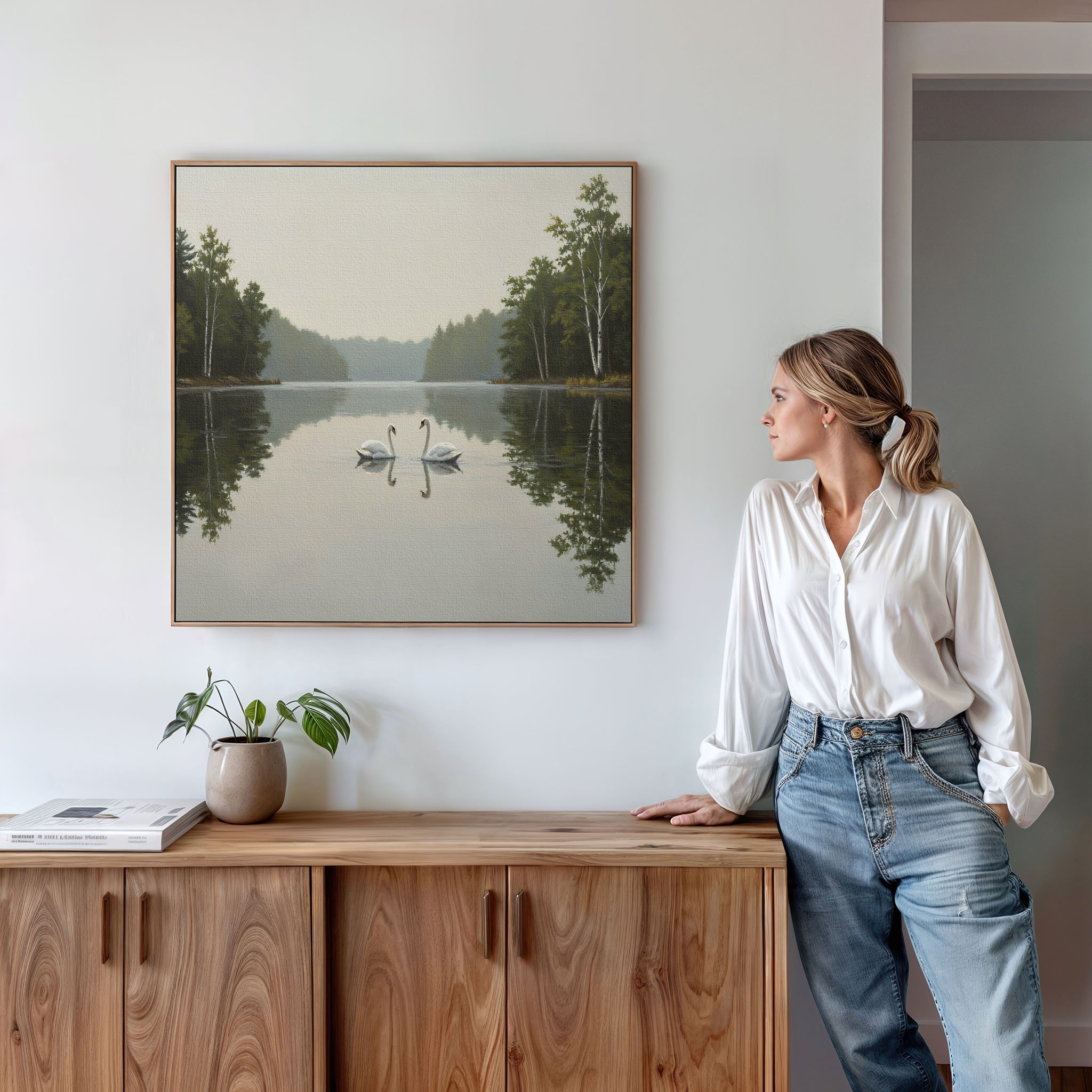 A woman in a white blouse and jeans stands by a wooden sideboard, admiring the Twin Swans Serenity Canvas Art - Square, which features serene swans with calm waters and trees, hanging on a white wall.