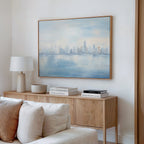 A minimalist living room features a wooden sideboard, stacked books, lamp, and bowl. The Urban Mirage Canvas Art - Horizontal depicting a misty city skyline hangs above. Cream-colored sofa cushions complete the modern look.