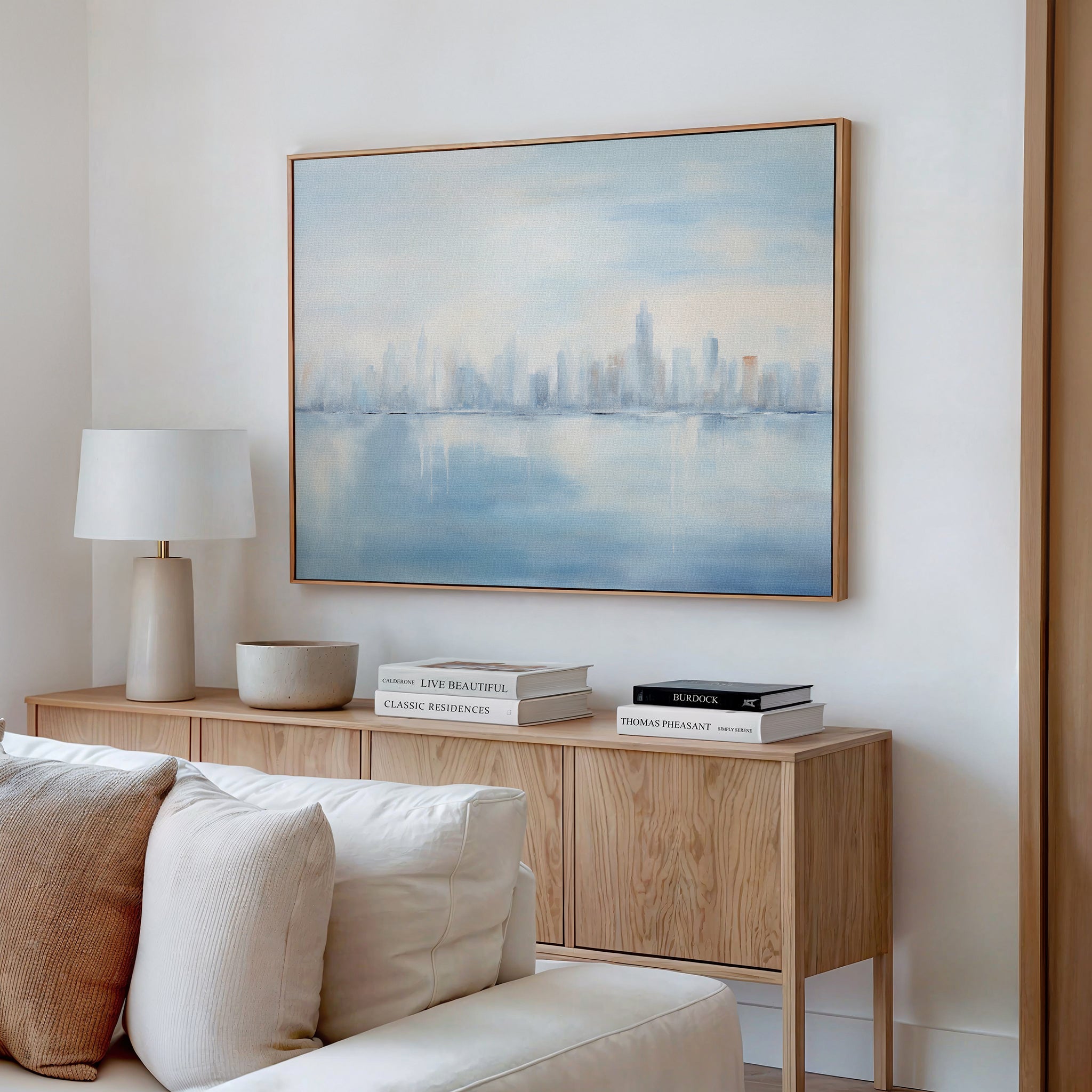 A minimalist living room features a wooden sideboard, stacked books, lamp, and bowl. The Urban Mirage Canvas Art - Horizontal depicting a misty city skyline hangs above. Cream-colored sofa cushions complete the modern look.