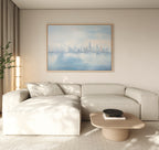 A modern living room with a light beige sectional, low wooden coffee table with black bowl, neutral walls, and the Urban Mirage Canvas Art - Horizontal featuring a soft blue city skyline above the sofa—perfect decor.