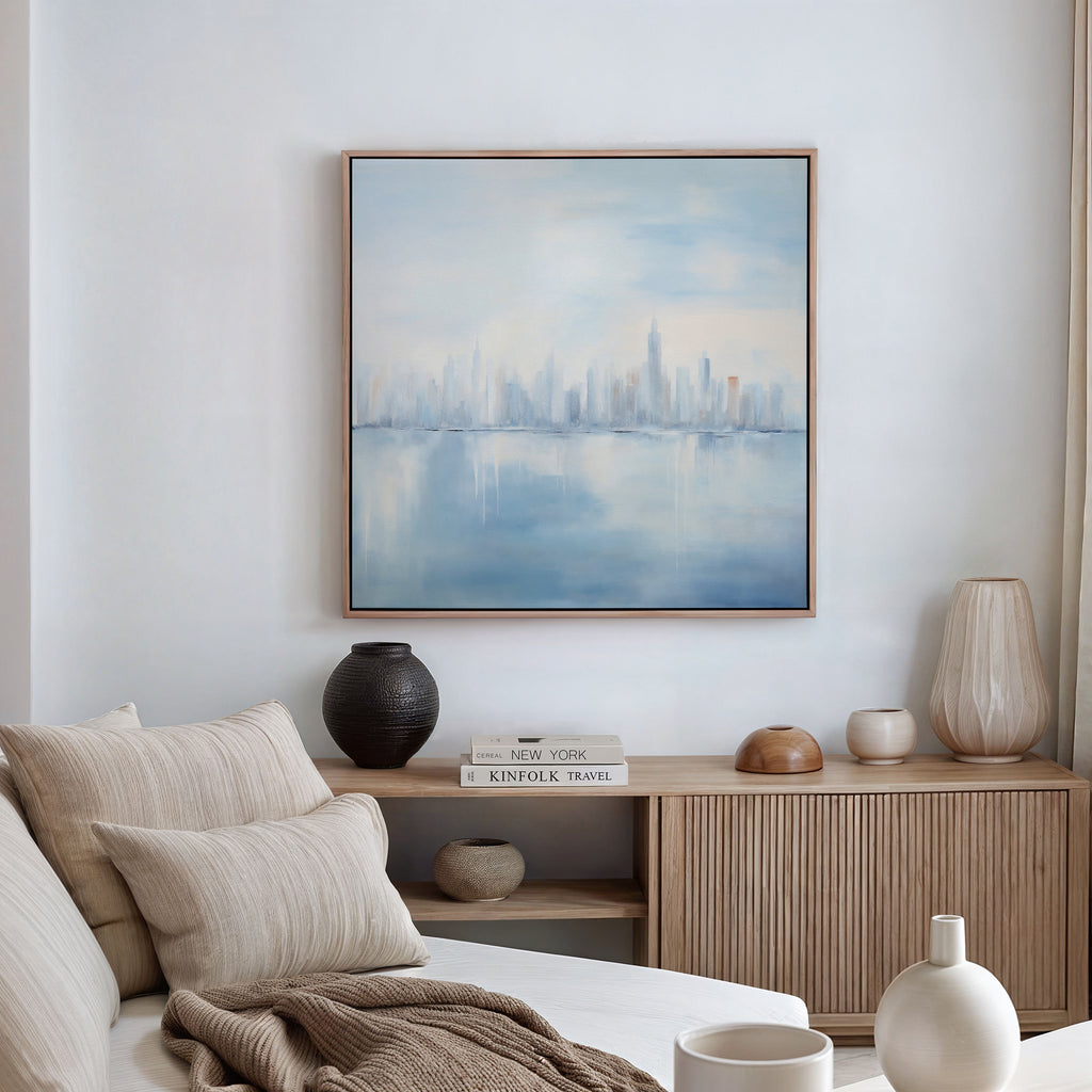 A serene living room features a beige sofa, neutral cushions, wooden sideboard, decorative vases, books, and the Urban Mirage Canvas Art - Square—a framed abstract painting of a city skyline reflected on water—highlighting the white wall.