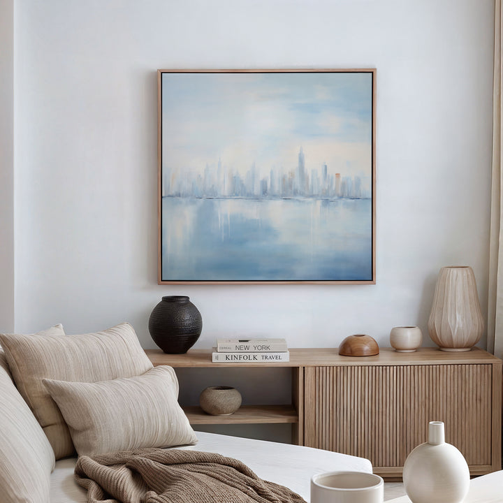 A serene living room features a beige sofa, neutral cushions, wooden sideboard, decorative vases, books, and the Urban Mirage Canvas Art - Square—a framed abstract painting of a city skyline reflected on water—highlighting the white wall.