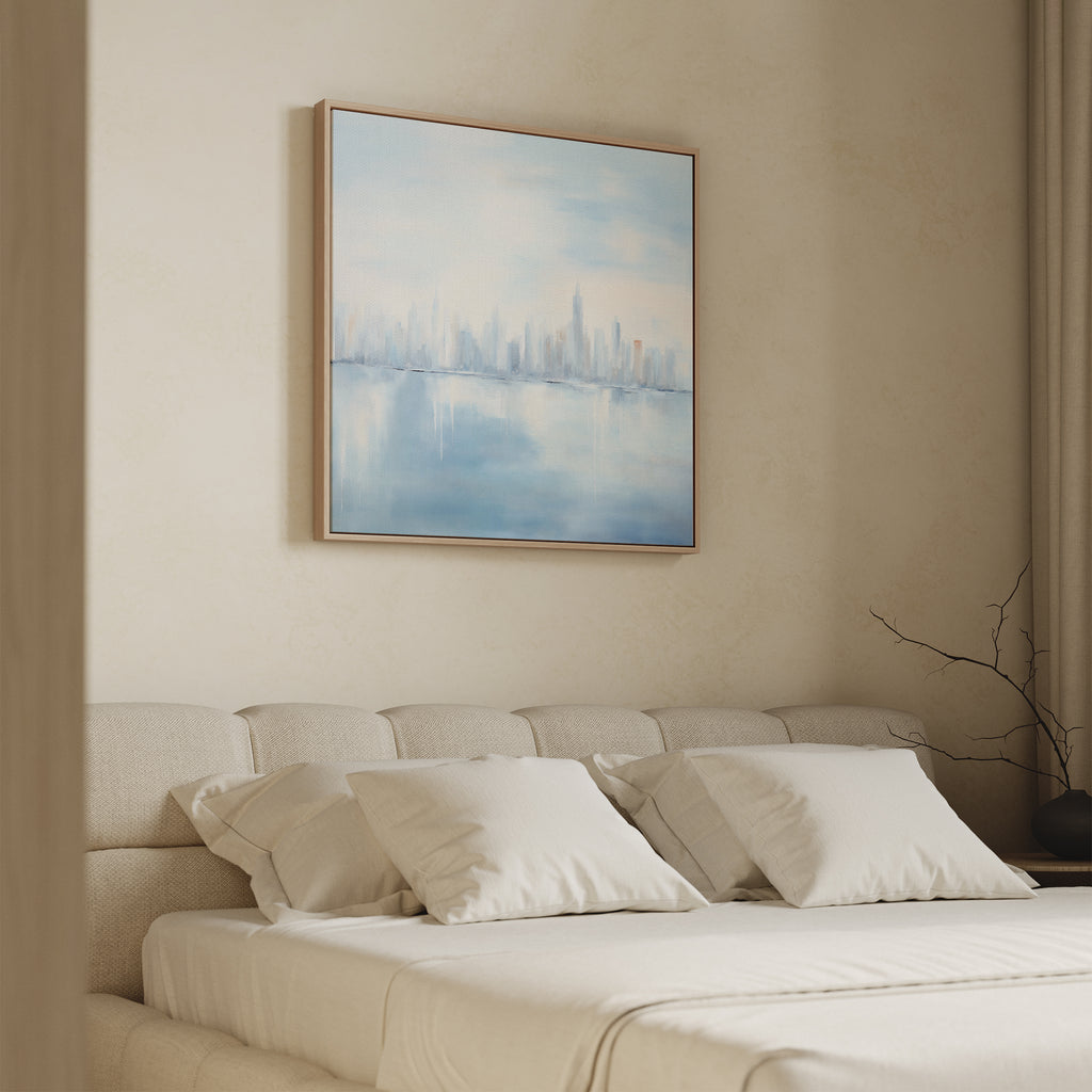 A beige bedroom features a neatly made bed with four white pillows and a light wood headboard. Above, the "Urban Mirage Canvas Art - Square" adds modern wall decor with a dreamy city skyline reflected on water.