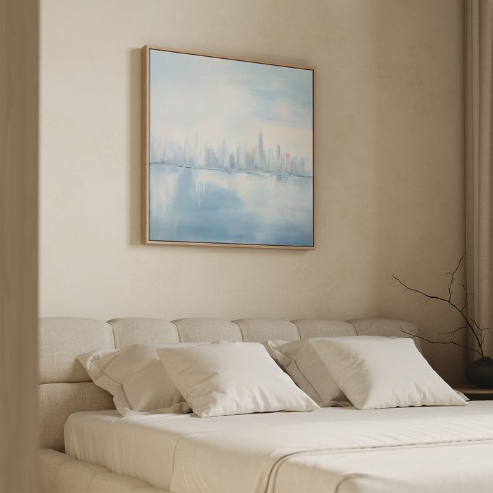 A beige bedroom features a neatly made bed with four white pillows and a light wood headboard. Above, the "Urban Mirage Canvas Art - Square" adds modern wall decor with a dreamy city skyline reflected on water.