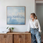 A woman in a white blouse and jeans leans on a wooden cabinet, admiring the Urban Mirage Canvas Art - Square with its dreamy city skyline. A potted plant and books complete the stylish modern living room decor.